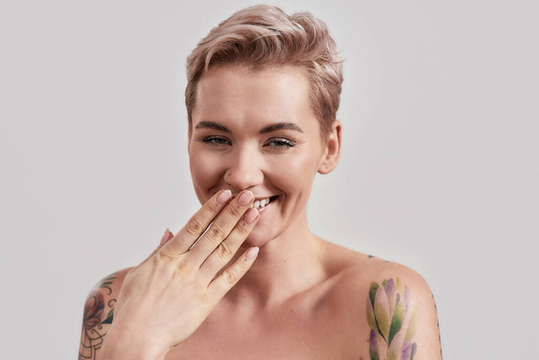 Funny. Portrait of a young attractive half naked tattooed woman with perfect skin laughing at camera, covering mouth with her hand isolated over light background - Foto, Imagem