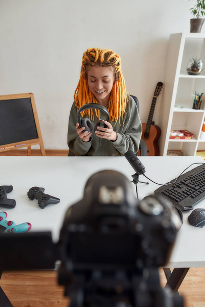 Female technology blogger with dreadlocks smiling, holding headphones while recording video blog or vlog about new gadgets at home - Фото, зображення