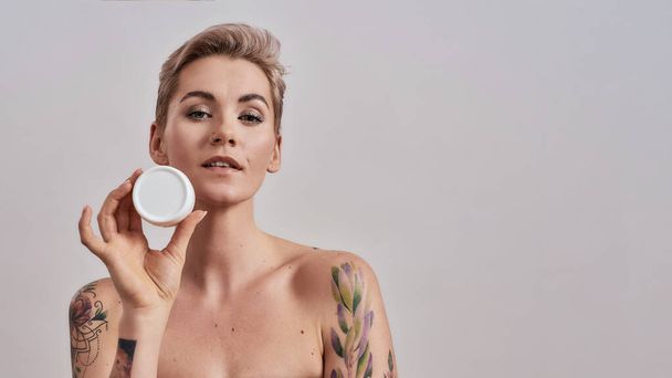 Beauty starts with skin. Portrait of beautiful tattooed woman with pierced nose and short hair holding white plastic jar of cream or body lotion isolated over grey background - Fotoğraf, Görsel