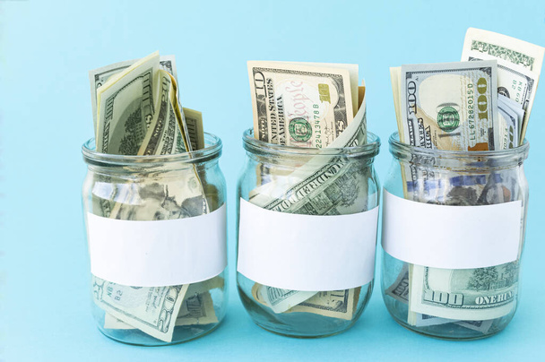 Three Piggy Bank from a Glass Jar for Life. New dollars sticking out of piggy banks on a blue background. The banks have white sheets for inscriptions. Mockup. Copy space. Economy concept. - Photo, Image