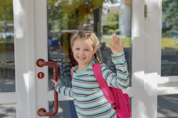 A girl with a pink satchel opens the school door. Children go out to study after vacation and the coronavirus epidemic. The new school year. - Foto, imagen