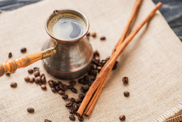 selective focus of cezve with coffee near cinnamon sticks and coffee beans on sackcloth - Фото, зображення