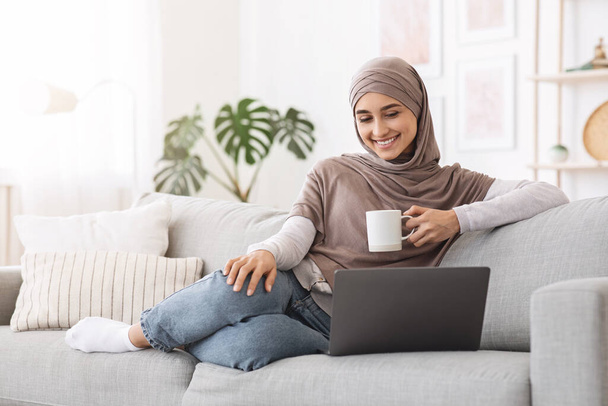 Passatempo em casa. Mulher árabe alegre assistindo filmes no laptop e bebendo café
 - Foto, Imagem