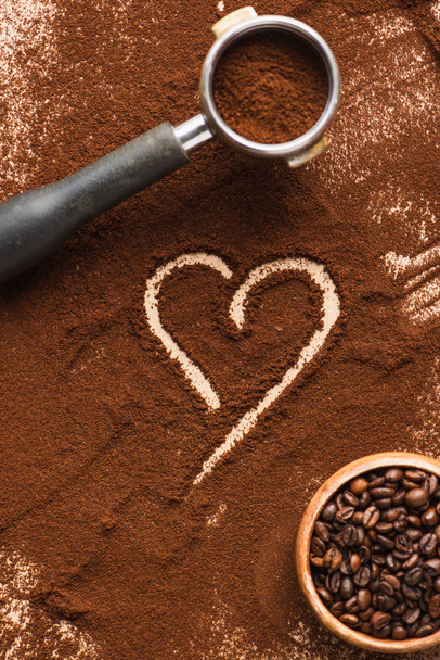 vista superior del corazón dibujado en el café molido cerca de frijoles en tazón y portafilter - Foto, Imagen