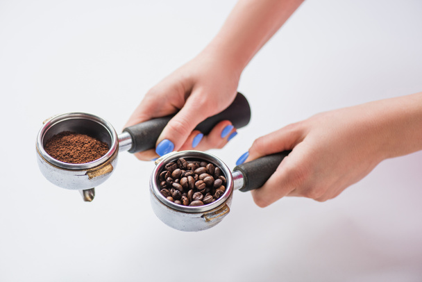 bijgesneden weergave van barista met portafilters met koffiebonen en gemalen koffie op witte achtergrond - Foto, afbeelding