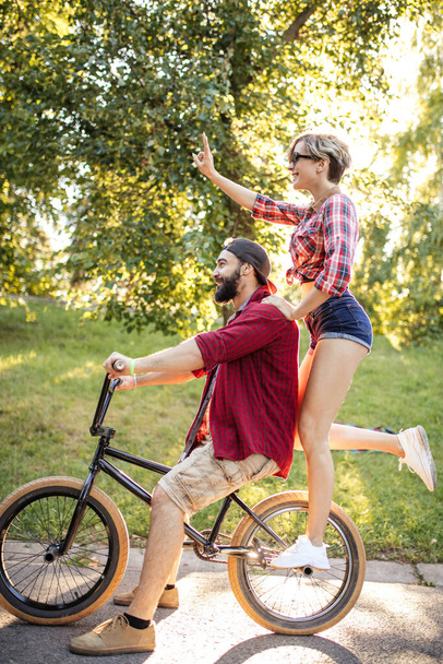 Ευτυχισμένο νεαρό ζευγάρι που πάει βόλτα με το ποδήλατο μια καλοκαιρινή μέρα στο πάρκο. - Φωτογραφία, εικόνα