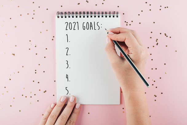 Hyvää uutta vuotta 2021. Naisen käsin kirjoittaminen 2020 Tavoitteet muistikirjaan koristeltu joulukoristeita tricolor tausta. Top view, tasainen lay - Valokuva, kuva