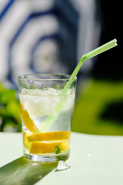 стакан с освежающим напитком со льдом, лимоном и соломой, на зеленом фоне и зонтиком. Летний напиток - Фото, изображение