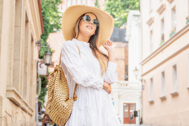 Young beautiful happy woman on vacation wearing summer hat and fashionable sunglasses. Tourist walking in the city street. - 写真・画像