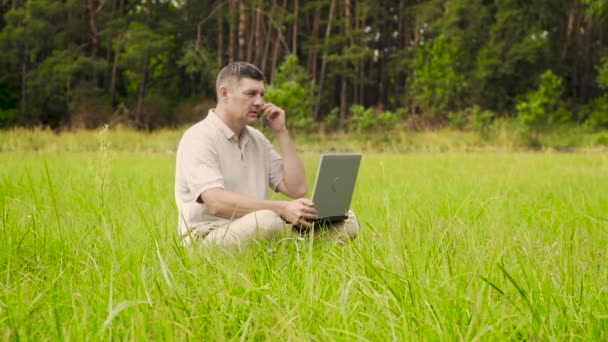 Ein Mann mit Laptop telefoniert auf dem grünen Rasen - Filmmaterial, Video