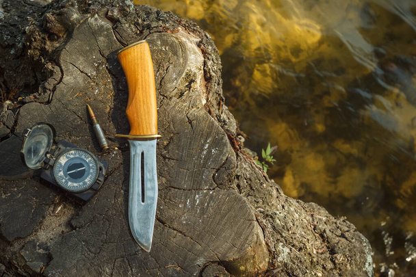 Αυτοδημιούργητο κυνηγετικό μαχαίρι, παλιά πυξίδα και φυσίγγιο 5.45 τοποθετούνται σε ένα κούτσουρο δίπλα στο νερό. Καλοκαιρινή σύνθεση με κυνηγετικό εξοπλισμό. Κοντινό πλάνο. - Φωτογραφία, εικόνα