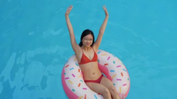 Samotna młoda azjatycka kobieta w czerwonym stroju kąpielowym pływająca w basenie w nadmuchiwanym okręgu różowy pączek podnosi ręce uśmiechając się patrząc w kamerę, strzelając z góry. Pochodzenie etniczne kobieta zrelaksować się w wodzie w kompleksie turystycznym - Materiał filmowy, wideo