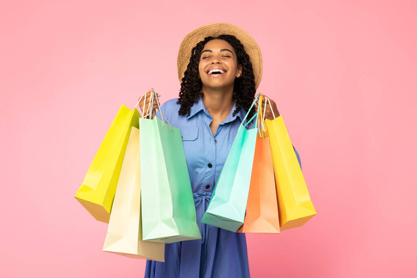 Chica negra riendo llevando bolsas de compras de pie sobre fondo rosa - Foto, imagen