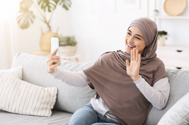 Video call. Muslim girl in hijab waving to smartphone camera at home - 写真・画像