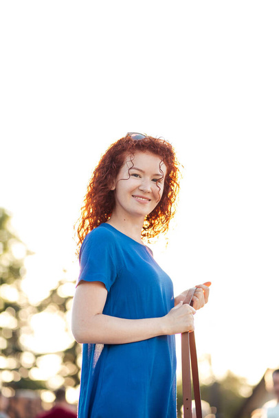 Young woman with waved curly red hair, in blue dress, in park. - 写真・画像