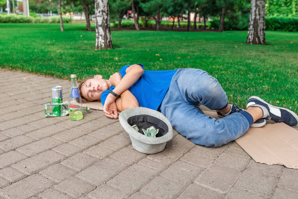 Пьяный бездомный подросток-нищий спит на улице, рядом бутылки с алкоголем. В кепке доллары. - Фото, изображение