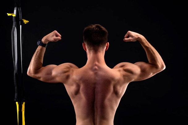Young bodybuilder with trx loops on a black background shows biceps, view from the back. Active lifestyle, sports. - Zdjęcie, obraz