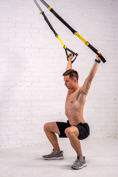 Muscular man doing exercises with trx straps, near a white brick wall. Bodybuilding. - Photo, image