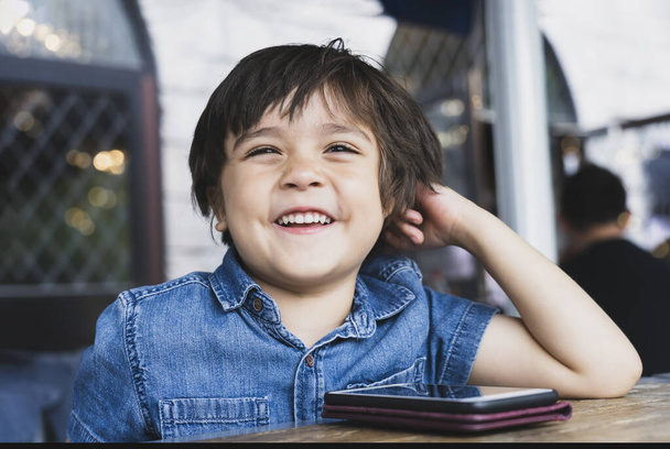 Портрет щаслива дитина, дивлячись з усміхненим обличчям, маленький хлопчик занадто дивиться або грає в ігри на мобільний телефон, чекаючи їжі в кафе, концепція "Діти з технологіями"
. - Фото, зображення