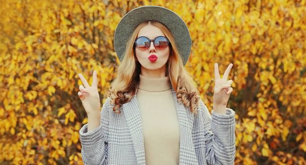 Retrato otoñal de mujer atractiva soplando labios rojos enviando beso de aire dulce usando abrigo gris, sombrero redondo sobre fondo de hojas amarillas - Foto, imagen