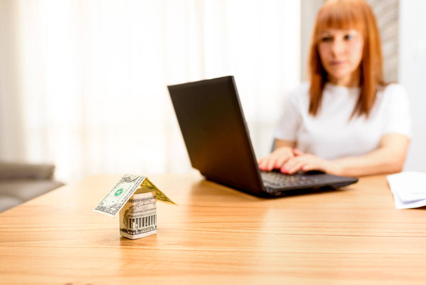Money house from dollar banknote on wooden table.Middle age worried woman using laptop sitting in the living room at home.Concept of buying a home, real estate activity, looking new homes for sale. - Photo, image