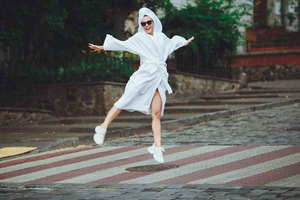 A glamorous lady in a white bathrobe with a white towel on her head and elegant sunglasses is walking along the street of a big city - Foto, Imagem