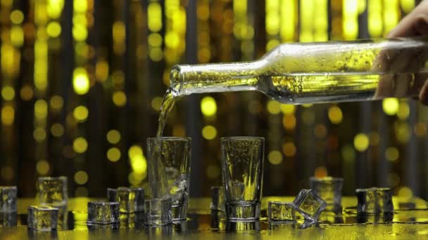 Barman vierta el vodka congelado de la botella en un vaso de chupito. Cubos de hielo contra fondo brillante fiesta de oro - Imágenes, Vídeo