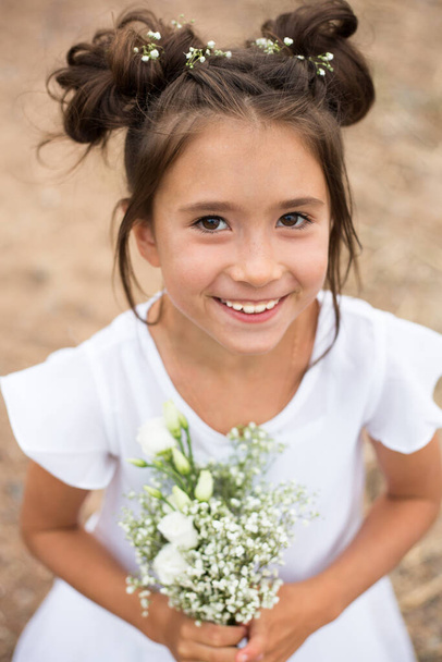una bambina tiene un mazzo di fiori bianchi su uno sfondo sfocato.capelli scuri, fiori bianchi nelle sue mani, vestito bianco - Foto, immagini