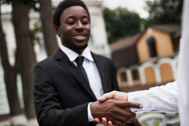 Στενή άποψη των δύο ανδρών επιχειρηματιών χειραψία. Συγκεντρώσου στους καρπούς. Νεαρός Αφρικανός Αμερικανός διευθυντής χαμογελά. Προσωπική συνάντηση συναδέλφων στο δρόμο. Πολυπολιτισμική εικόνα. - Φωτογραφία, εικόνα