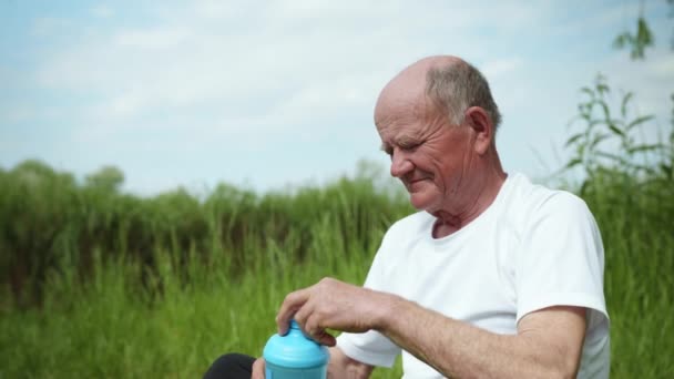健康的な高齢者の男性は、オープンエアのマットに座ってスポーツや瞑想をした後、スポーツ栄養のためのボトルからバランスを回復し、きれいな水を飲む - 映像、動画