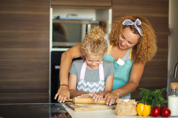 Beautiful young mom and her cute little daughter rolling out dough at home. - Foto, Bild