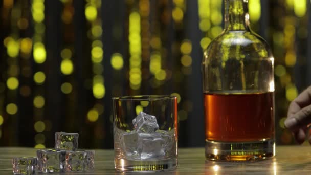 Nalití zlaté whisky, koňaku nebo brandy z láhve do sklenice s kostkami ledu. Lesklé pozadí - Záběry, video
