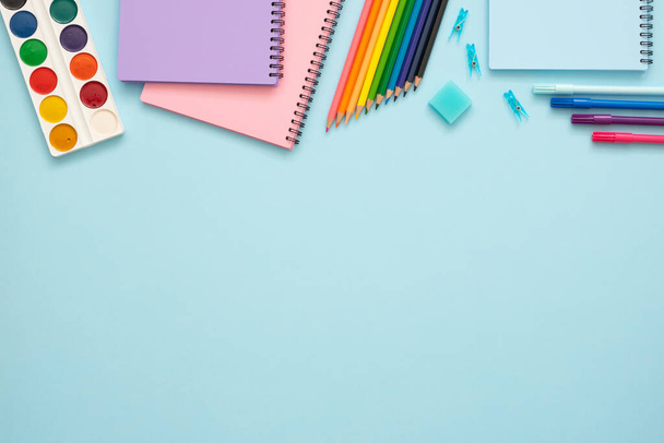 Education concept. Colorful stationery set on blue background. Top view of blue, pink and purple notebooks, watercolor, paper pins, eraser, felt tip pens and color pencils. Copy space.  - Fotó, kép