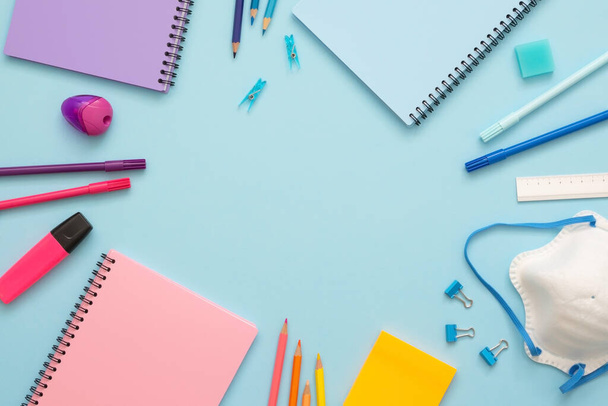 Colorful stationery set on blue background. Top view of blue, pink and purple notebooks, paper clips, eraser, felt tip pens, color pencils, marker and face mask. Copy space. Back to school. - Photo, image