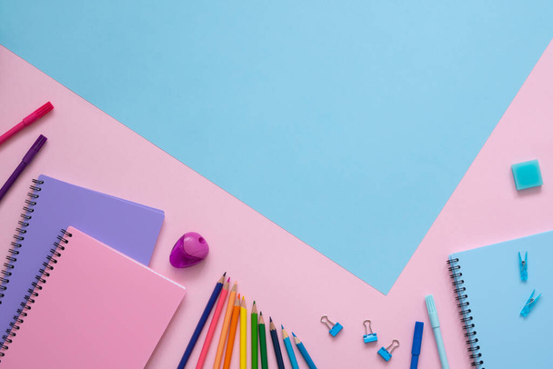 Back to school. Blue, purple and pink notebooks, color pencils, felt tip pens, pencil sharpener, eraser and paper clips on pink background and copy space on blue background. Top view. - Photo, image