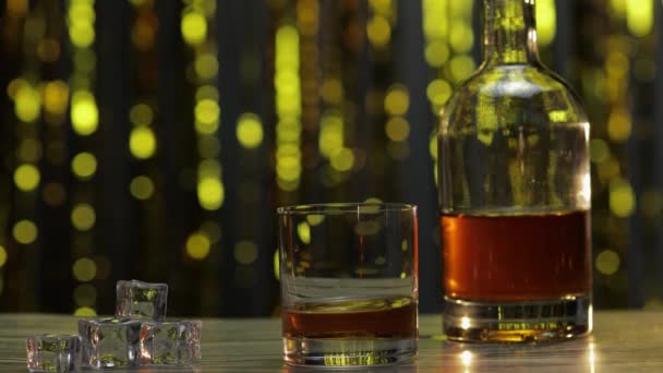 Baarimikko lisää jääpaloja lasiin, jossa on kultainen viski, konjakki tai brandy pöydällä. Kiiltävä tausta - Materiaali, video