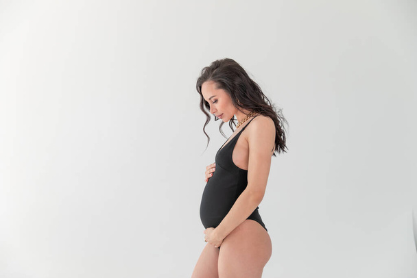A fashion stylish pregnant sexual woman in Vogue style. beautiful Girl with belly for 9 months waiting for baby. On a white gray background. glamorous Pregnant in a black bodiesuit. Copy space - Fotó, kép