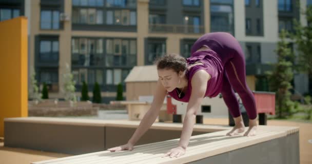 Nő csinál nyújtás és jóga gyakorlatok a padon az udvaron egy apartman épület, fitness a városi környezetben, jóga osztály szabadban, fizikai edzés a városban, 4k 120p Prores HQ - Felvétel, videó
