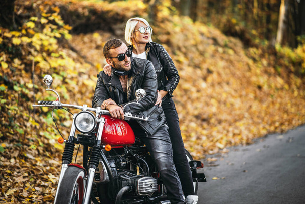 Pretty couple near red motorcycle on the road in the forest with colorful blured background. Relationship concept. - Φωτογραφία, εικόνα