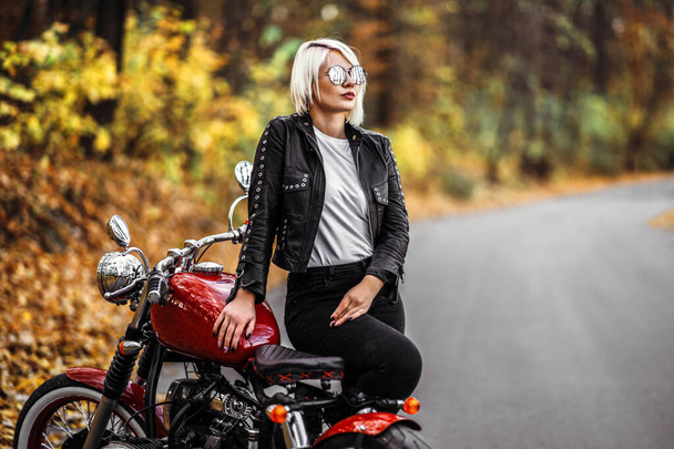 Pretty blonde biker girl in sunglasses with red motorcycle on the road in the forest with colorful blurred background. - Fotó, kép