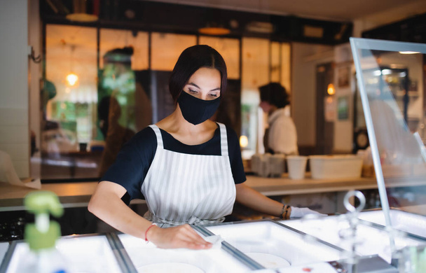 Young woman with face mask working indoors in cafe, disinfecting surfaces. - Foto, Bild