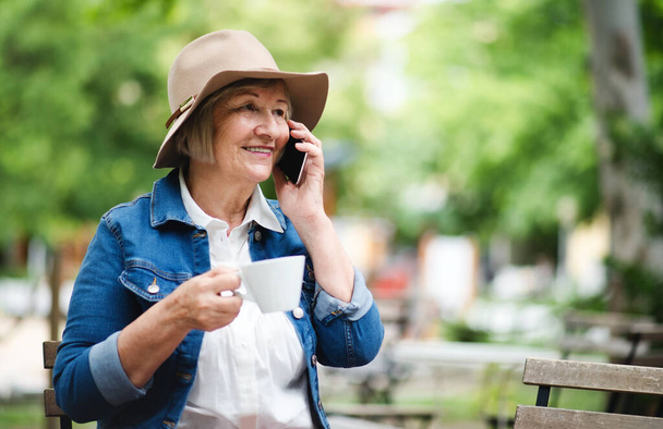 Senior woman with hat sitting outdoors in cafe, using smartphone. - Valokuva, kuva