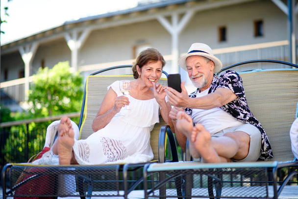 Senior couple with wine and smartphone outdoors on holiday, relaxing. - Photo, image