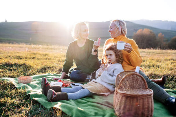 Annesi ve büyükannesi olan küçük bir kız gün batımında doğada piknik yapıyor.. - Fotoğraf, Görsel