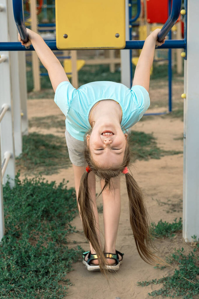 Милая маленькая девочка с двумя смешными хвостами висит вверх ногами и веселится на детской площадке. - Фото, изображение