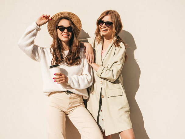 Two young beautiful smiling hipster girls in trendy white sweater and coat. Sexy carefree women posing near wall in the street in hat and sunglasses. Positive models having fun and drinking coffee  - Фото, изображение