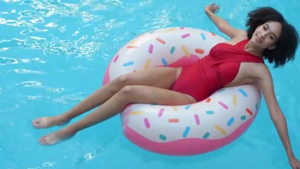 カメラで笑みを浮かべてピンクのドーナツの上から撮影した後、ホテルのホームプールで休憩中の巻き女性日焼け。アフロティーン女の子で赤水着楽しいです青水とともにインフレータブルリングアップ - 映像、動画