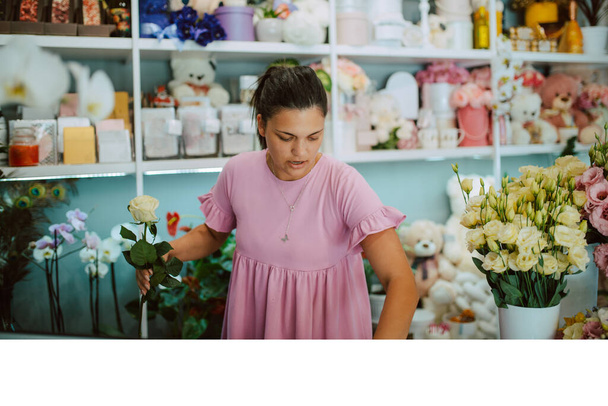 Young attractive brunette caucasian woman florist in a pink dress arranging a bouquet of flowers in a flower shop - 写真・画像