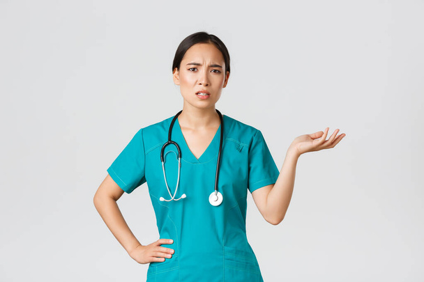 Covid-19, healthcare workers, pandemic concept. Frustrated and annoyed asian female doctor, nurse in scrubs raise one hand and looking questioned and pissed-off, complaining something - Photo, image