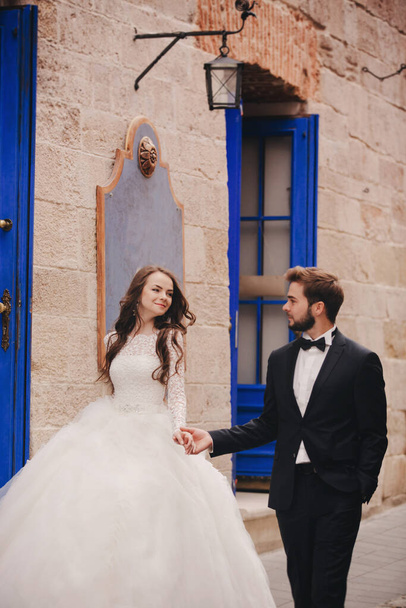 Esküvői pár ölelkezik az óvárosban. Kék évjárat ajtók és kávézó az ősi városban a háttérben. stílusos menyasszony fehér hosszú ruha és vőlegény öltöny és csokornyakkendő. esküvő napja. - Fotó, kép